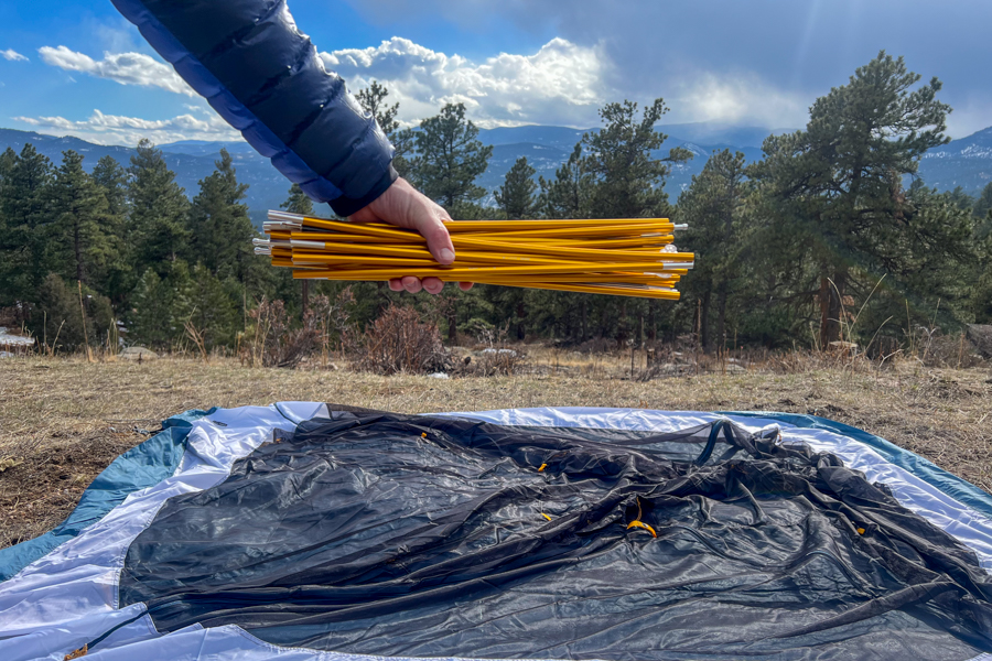 A hand holds gold colored tent poles up to the camera. A tent body is layed out on the ground. There are pine trees and rain clouds in the distance.