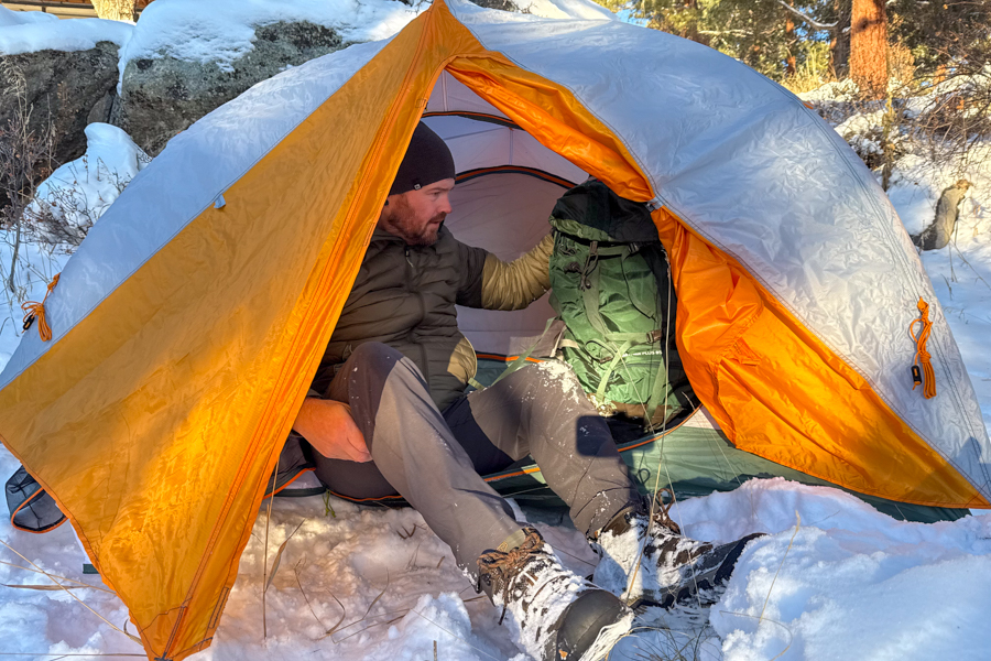 A person sitting at the open doorway of the tent in snowy conditions, pulling a large backpack into the vestibule while wearing winter boots and a warm jacket.