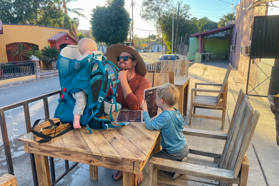 Osprey Poco SLT carrier set on a wooden table outdoors, with a baby seated inside and a family enjoying their meal in a casual setting.