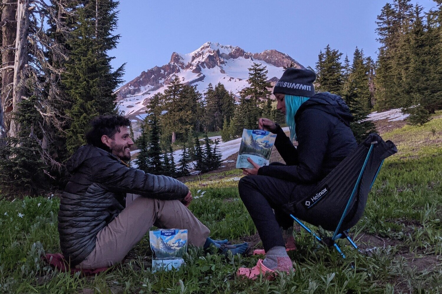 Two hikers eating Backpacker's Pantry freeze-dried meals in front of a mountain